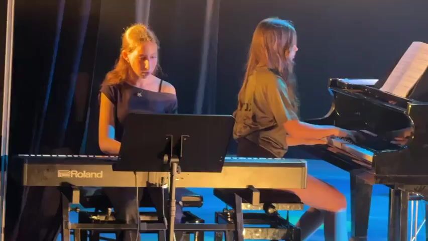 Two young girls are playing pianos on a stage. The girl on the left is playing a Roland keyboard and the girl on the right is playing a grand piano. They are both focused on their music. The stage is lit with blue and green lights. There are dark curtains behind the pianos.