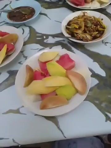 A close-up, slightly shaky video shows a table laden with several plates of cut fruit and dipping sauces. There are at least three plates of sliced fruit, including watermelon, mango, and guava. One plate contains a mixture of what appears to be savory salad with vegetables and possibly peanuts. Two small bowls of dark, likely savory, dipping sauce are also visible. The hands of at least two people are visible interacting with the food, one person is using a skewer to pick up a piece of fruit and dip it into a sauce, while another person is holding a piece of fruit on a skewer to their mouth. The table has a marbled, abstract pattern in shades of white, gray, and gold. The background is out of focus but suggests an indoor or covered outdoor setting. The overall impression is of a casual meal or snack.