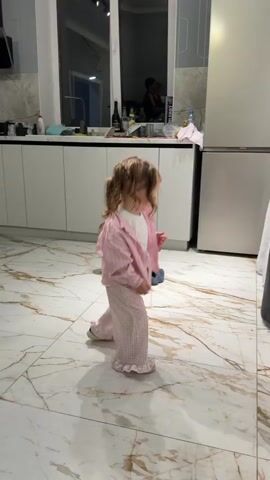 Two toddlers are in a kitchen with white cabinets and beige-marbled floors. One toddler, a fair-skinned baby with light hair, is crawling on the floor towards the left. The other toddler is a light-skinned girl with blonde, curly hair tied into pigtails. She is wearing a pink striped shirt and pink plaid pants with ruffles. She walks and spins erratically in place. Pop music plays in the background.