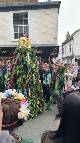 A crowd of people watch a procession in a European-looking town. A figure covered in green leaves with colorful ribbons and a crown on top moves through the crowd. The crowd is largely made up of adults, though there are some children. Some are dressed in green, some wear flower crowns, and others are holding up phones to record the event. The people stand on cobblestone streets, and buildings line the street.  Confetti flies through the air. A shop sign that says "Through the looking glass" can be seen.