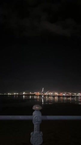 The video captures a nighttime scene from a pier or similar elevated structure. A metal railing, painted a light blue-gray, is visible in the foreground. Beyond the railing, the water reflects the lights of a distant cityscape. The city lights are a mix of white, yellow, and red, creating a blurred reflection on the water's surface. The sky above is dark, with some cloud cover visible. The overall scene is dark and relies on the city lights for illumination.