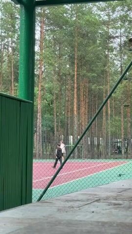 This video captures an outdoor scene on a cloudy day, likely in the late afternoon or early evening, judging by the diffused light. The setting appears to be a sports court, possibly for tennis or volleyball, surrounded by a dense forest of tall pine trees. The court itself has a reddish-pink and green surface, divided by a white line.

In the foreground, a green fence and a partially visible wooden structure suggest a viewing area or a shaded spot. Beyond the fence, a net encloses the court.

Three people are visible on the court. Two individuals, dressed in dark clothing, are in motion. One person in a black t-shirt and shorts is running towards the right of the frame, while another, also in dark attire, is walking. A third person, wearing a white hoodie and dark pants, is standing on the court, seemingly engaged in a game or activity. Their postures suggest an informal gathering, perhaps playing a sport or simply enjoying some outdoor time together.

The presence of tall pine trees indicates a natural, possibly recreational area. The overcast sky suggests mild weather, not bright sunshine. The overall atmosphere seems relaxed and casual. There is no visible text in the frame. The location context of Samara, Russia, places this scene within a temperate climate known for its forests.