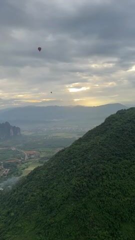 The video captures a scenic aerial view of a valley with rolling green hills and mountains in the distance. Two hot air balloons are visible in the sky. One balloon is closer and appears red, while the other is smaller and further away. The sky is partly cloudy with sunlight breaking through, casting a warm glow over the landscape. A lush green mountainside dominates the foreground. The valley below is dotted with buildings, agricultural fields, and a road or path. The overall impression is one of natural beauty and a peaceful morning in a mountainous region.