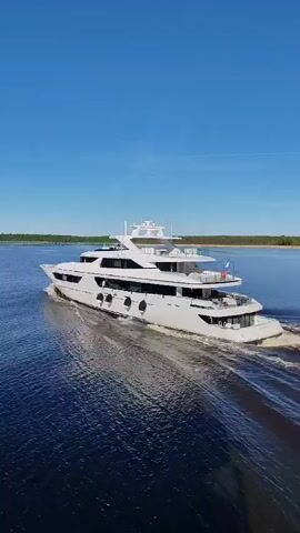 A white superyacht is seen from a high angle, moving away from the viewer on a body of water under a clear blue sky. The yacht has multiple decks and is leaving a wake behind it. The water is dark blue, and there are trees visible on the distant horizon on both sides of the water. The sun is shining brightly, creating reflections on the water's surface.