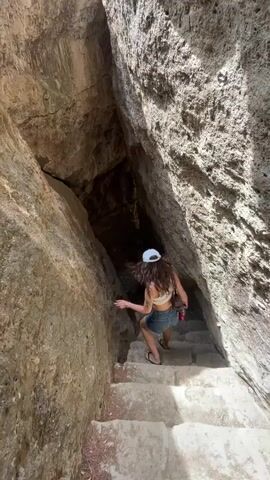 A person wearing a white baseball cap, a light-colored crop top, and denim shorts walks up a stone staircase that leads into a narrow opening between two large rock formations. The person's back is to the camera, and they are holding a red object in their right hand. The rock formations are rough and textured, and sunlight illuminates the scene, casting shadows. The staircase appears to be carved into the rock or made of large, irregular stone blocks. The opening into which the person is walking is dark.