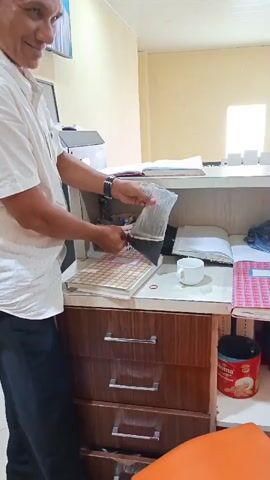 A person in a white shirt and black pants is standing at a wooden desk with drawers. They are holding a clear plastic bag with black contents, which they are tearing open. On the desk are several notebooks, a white mug, and a red rubber band. To the right of the desk, there is a shelf with more notebooks and a red tin can with a picture of cookies on it. The environment appears to be an office or room with pale yellow walls and a white ceiling.