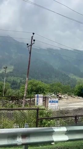 The video shows a mountain landscape under a cloudy sky. In the foreground, there is a road and some construction materials. A fence runs along the left side of the road. On the right side of the road, there are several large rocks and piles of dirt. There are also two white boards with blue lettering that appear to be signs for "ARCO CEMENT". A person in military uniform is standing near the construction materials. The background consists of green trees and the slopes of mountains. The camera pans slowly from left to right.