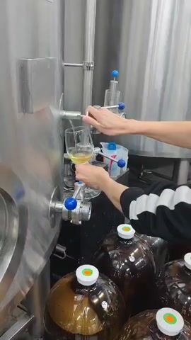 A person's hands are seen operating a valve on a large, stainless steel tank, filling a glass with a pale yellow liquid. Below the tank, several large, brown plastic carboys with green and white caps are visible. The environment appears to be an industrial or production facility, possibly a brewery or winery, with shiny metal surfaces and equipment.