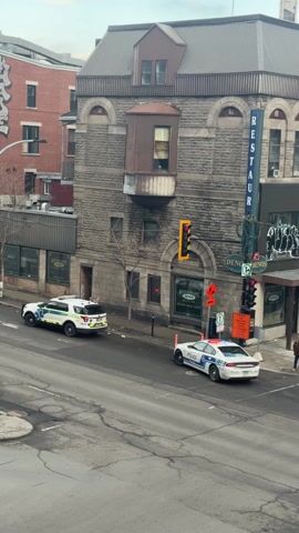 The scene captured is an urban street corner on an overcast day. A distinctive gray stone building, with a bay window, dominates the right side of the frame and features a vertical "RESTAURANT" sign. Two police vehicles, one a white SUV-type and the other a white sedan, are positioned on the street, with the SUV on the left side of the frame and the sedan angled on the right. A silver car with a roof rack approaches the intersection from the lower left. Pedestrians are visible near the restaurant. Traffic signals at the corner show a yellow light. The overall atmosphere suggests a moment of observation or potential action, with the presence of law enforcement setting a serious tone.