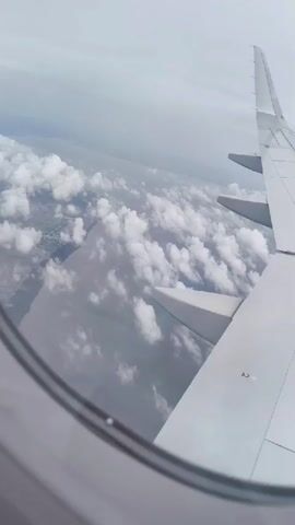 The video is a first-person view from an airplane window. The viewer is looking out at a cloudy sky and the wing of the airplane. The clouds are white and fluffy, and they are spread out across the sky. The wing is white and has some markings on it. The land below is not clearly visible due to the clouds. The video is shaky, suggesting it was filmed by someone holding a phone.