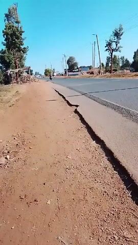 The video shows a road in a rural or semi-rural area on a sunny day. The road appears to be paved, but the shoulder is cracked and broken. There are utility poles and lines visible along the roadside. On the side of the road, there are buildings, some with clotheslines and laundry hanging outside. A few cars drive on the road and a person wearing a jersey with the number 6 on the back is walking on the cracked shoulder of the road.