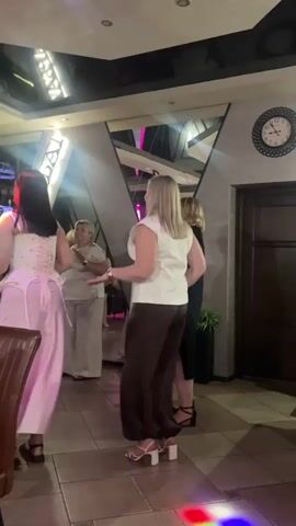 This media file captures a lively indoor scene, likely a celebration or social gathering, possibly a wedding reception or a birthday party, taking place in Orsha, Belarus. The focal point is a group of women, primarily in the foreground and mid-ground, engaging in dancing or mingling.

On the left, a woman with dark hair is adorned in a pink corset-style top and a flowing pink skirt, her posture suggesting movement and enjoyment. Adjacent to her, an older woman in a light-colored outfit stands with her hands clasped, observing the scene. To the right, a blonde woman in a white sleeveless top and dark wide-legged pants, and another woman in a black dress, are also captured in the midst of the event. The presence of a clock on the wall indicates that the event is taking place during daytime or early evening.

The setting appears to be a well-decorated indoor venue, possibly a restaurant or a banquet hall. The ceiling features modern lighting fixtures, and a large mirrored wall with a geometric design adds a touch of sophistication. The floor is tiled, and the overall ambiance is enhanced by dynamic colored lighting that illuminates the space and the dancers, creating a festive atmosphere. The lighting shifts from green and blue to pink and red, suggesting the presence of disco lights or stage lighting, indicative of entertainment and celebration. A chair in the foreground suggests a seating area for guests.

Visible text is limited but a sign with the Cyrillic letters "АРБА" (ARBA) is partially illuminated on a structure in the background, possibly a stage decoration or a name of the venue. The overall mood is joyful and energetic, with the people appearing engaged and happy, caught in moments of dancing and socializing.