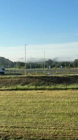 The video displays a rural landscape with a clear blue sky and some distant mist. In the foreground, there are fields of grass and crops, with a dirt path winding through them. A simple house with a white facade and a red roof is visible in the middle ground, surrounded by trees and a small concrete structure. In the background, a highway with a white semi-truck and several other trucks parked can be seen. There are also power lines and streetlights along the highway. The overall impression is of a tranquil morning in the countryside.