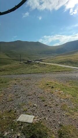 The video begins with a close-up shot of the underside of a green structure, possibly a cable car cabin, with its wheels and cables visible against a blue sky with scattered clouds. The camera then pans to reveal a mountainous landscape with rolling green hills, a dirt road, and a small body of water. As the camera moves, the view shifts to show more of the mountain range, with patches of dry grass and rocky terrain in the foreground. The video appears to be shot from a moving vehicle, likely the cable car itself, as evidenced by the changing perspective and the presence of the cabin's structure. There are no people or distinct interactions visible. The environment is natural and scenic, suggesting a mountainous region during daytime.