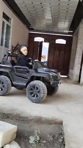 A toddler in a black hooded jacket and striped hat sits in a black toy Jeep, positioned on a concrete surface. The vehicle features large, knobby tires and off-road lights. The toddler maneuvers the car, bumping into a rusty metal pole attached to the side of a light-colored house. Behind the vehicle, a gate opens to reveal a parked car in the distance. The area above the toddler is covered by a metal awning, and a soccer ball rests near the house.
