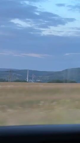 The video shows a view from a moving vehicle, looking out at a landscape. The sky is a deep blue with some clouds. In the distance, there are rolling hills covered in trees. In the foreground, there is a field of tall grass that appears blurred due to the motion of the vehicle. There are also power lines and a tall industrial structure visible in the distance. The overall impression is one of a rural landscape during twilight.