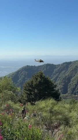 A yellow helicopter flies over a lush green mountainside under a clear blue sky. In the foreground, people are visible on a path surrounded by green foliage and pink flowers. The helicopter is in mid-flight, and its rotors are spinning. The mountains are densely covered in trees, and in the distance, a hazy landscape can be seen. The lighting suggests it is daytime.
