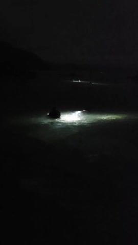The video shows two people in a dark, shallow body of water, possibly a sea or ocean, at night. Each person has a bright light, illuminating the water around them. One person is closer to the camera, kneeling or crouching in the water, with their light pointing forward. The other person is further away, also in the water, with their light directed towards the surface. The environment appears to be a dark shore, possibly with hills or land visible in the background. The focus is on the illuminated areas in the water created by the two lights.