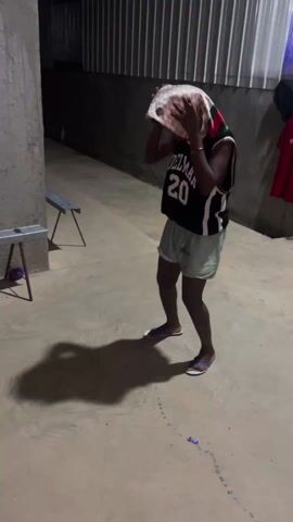 A child dances to music in an outdoor, paved area. The child, who has dark skin, wears a white-patterned headscarf, a black and white basketball jersey with “DEL MAR 20” printed on it, and light green shorts. The child wears flip flops and is facing the camera, moving their arms and legs to the music. The area is dimly lit, casting a long shadow of the child on the ground. To the left of the child are two metal benches. Behind the child, there is a wall of corrugated metal and two items of clothing hanging up.