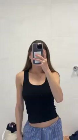 A young woman stands in a bathroom and takes a selfie in the mirror. She is wearing a black tank top and blue and white striped pajama pants. Her face is obscured by her phone. She makes a peace sign with her left hand. The bathroom has white tiled walls. A chrome towel rack is visible on the wall.