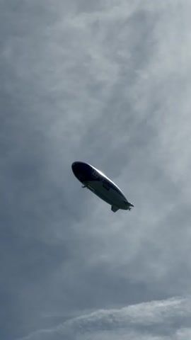 The video shows a blimp flying in the sky. The blimp is white and blue and has writing on its side. The sky is cloudy and the blimp is tilted at an angle. The video is shot from below.