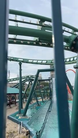The video shows a water ride at an amusement park. A roller coaster track, painted green, curves above a water channel. Boats, with passengers visible, are moving through the water. The track has lights on it, and supports are visible. In the background, buildings and a cloudy sky are present. The video appears to be shot from behind a fence.