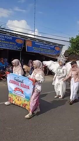 The video shows a parade, presumably a cultural event, taking place on a street. Several people in elaborate costumes walk in the procession. There are young women dressed in traditional clothing and hijabs, carrying a banner that reads "FESTIVAL NAVAL BUDAYA." 

One individual is adorned with large white wings and an ornate headpiece, resembling an angel. A woman in a pink jacket and white skirt is walking beside the "angel", holding her hand. Following them is a person wearing an elaborate mask and costume, with a long orange train. The street is lined with spectators, including people of all ages.

Shops with signs written in a language other than English are visible in the background. The sky is blue, and the weather appears to be sunny. Overall, the video captures a lively and colorful cultural celebration.