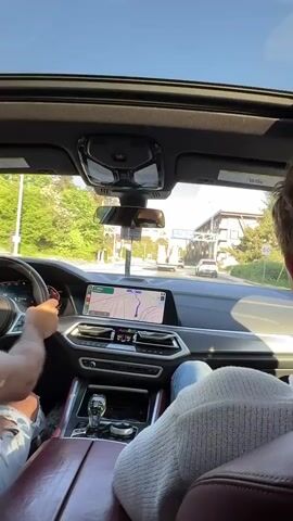 A car is driving into the city of Nanaimo, Washington. The driver has a passenger. The dashboard in the car contains a GPS map. There is an open sunroof. The car passes a truck with several large objects wrapped in orange tape.