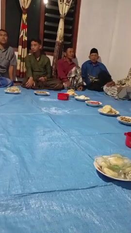 The video shows a gathering of at least six men sitting on a blue tarp spread on the floor. They appear to be having a meal together, as there are several plates of food and some red bowls arranged on the tarp in front of them. The men are dressed in casual clothing, with some wearing traditional Islamic headwear. The setting appears to be indoors, possibly a home, with a window visible in the background. The lighting is dim, suggesting it might be evening or nighttime. The men are interacting with each other, with some appearing to be engaged in conversation. The overall atmosphere seems relaxed and social.