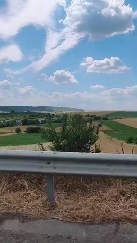 The video shows a view from a moving vehicle looking out over a rural landscape. The foreground features a metal guardrail and dry grass. Beyond the guardrail, fields of varying colors, likely crops, are visible, arranged in a patchwork pattern. Some fields are green, while others appear to be harvested or in a different stage of growth, displaying lighter, straw-like colors. Trees and scattered buildings dot the landscape in the distance. The sky is a bright blue with fluffy white clouds. The overall impression is one of a sunny day in the countryside.