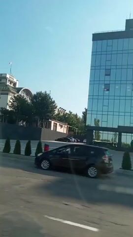 This media file captures a moving view of a street scene in Băcioi, Moldova. The time of day appears to be daytime, with a clear blue sky suggesting fair weather.

The primary subjects are vehicles and buildings. In the foreground, a silver car with "Letz taxi app" and "VISA" branding is visible, along with a person walking behind it. Further down the street, a light blue bus with the number "818" displayed prominently on its side is passing by. The bus has a graphic on its side featuring a white vehicle.

The background features several buildings. One is a modern, multi-story glass-fronted building. Another is a more ornate building with the text "PRESIDENT HOTEL" visible. Trees and greenery are abundant throughout the scene, indicating a potentially suburban or park-like setting. The overall impression is that of a typical day in a town or city, with traffic and commercial activity.