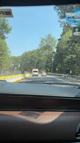 The video, filmed from inside a car, shows a daytime scene on a winding road, likely a highway, in Coajomulco, Mexico. The weather is clear and sunny, with a blue sky visible above.

The view is from the driver's or passenger's perspective, looking through the front windshield over a dark dashboard with a brown lower panel. On the upper right of the windshield, an "MX" sticker is visible.

Ahead, a white van with green and white markings on its rear is driving, followed by a silver sedan. Other vehicles can be seen in the distance. The road has multiple lanes and features yellow and grey concrete barriers on the left. Yellow chevron road signs with black arrows, indicating curves, are visible on both sides of the road.

The road is flanked by dense green trees, primarily conifers, suggesting a mountainous or heavily forested area. The audio contains Spanish speech: "Te acuerdas de la..." (Do you remember the...).