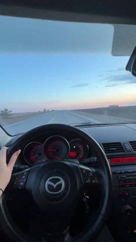 The video shows a first-person perspective of someone driving a Mazda car on an open highway. The sky is visible through the windshield, showing a sunset with hues of blue and orange. The car's dashboard is visible, with the Mazda logo on the steering wheel and red lighting on the console. There is a water bottle in the passenger's side door. The road appears straight and relatively empty.