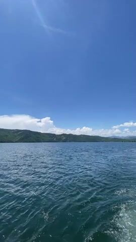 This is a vertical, first-person perspective video of a body of water under a bright blue sky with scattered white clouds. In the distance, a line of green, tree-covered mountains stretches across the horizon. The camera appears to be on a moving watercraft, as indicated by the wake visible in the lower right corner of the frame. The water is choppy and reflects the sunlight. The overall impression is of a calm, sunny day on a large lake or bay.