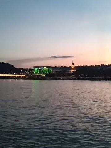 The video shows a wide shot of a river with a city skyline in the background during sunset. The sky is painted with hues of pink, purple, and blue. A modern building with green lights illuminates the left side of the skyline, contrasting with a traditional church steeple on the right. The water is calm with gentle ripples. The overall scene evokes a peaceful and serene evening ambiance.