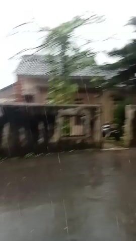 The video shows a street scene during heavy rain. The street is flooded and buildings are visible in the background. A person walks across the street in front of a house. The camera appears to be looking out from under a sheltered area. The lighting is dim due to the overcast sky.