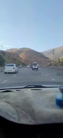 The video shows a view from inside a car traveling on a highway. The highway is surrounded by mountains. Several cars and vans are visible on the road, all traveling in the same direction. The sky is clear and blue. There is a car air freshener on the dashboard in the foreground.