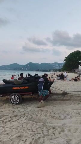 The video shows several people on a sandy beach beside the ocean on an overcast day. Mountains can be seen in the background. The main focus of the scene is a black Sea-Doo jet ski on a small trailer on the beach. Two men are standing near the jet ski, one of whom is holding a rope attached to the trailer. Further down the beach, a row of three other jet skis on trailers are parked, with their front ends facing the ocean. People are scattered along the beach, some standing near the water, some swimming, and some relaxing on the sand. There are also some trees and structures that could be beach huts or umbrellas.