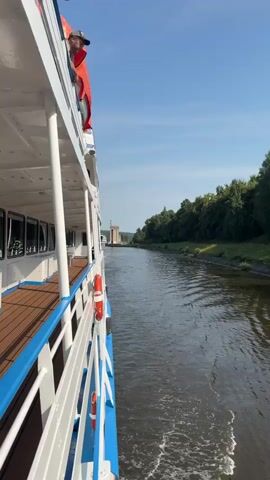 The video begins with a side view of a white passenger boat with blue trim, moving along a canal. A person wearing a red shirt and a cap is visible on the upper deck of the boat, looking out towards the water. The boat is on the left side of the frame, and the canal is to the right, with lush green trees lining the banks. As the video progresses, the camera shifts focus towards the right, revealing a large, light-colored building on a concrete platform in the middle of the canal. The building has a classical architectural style with columns and a flat roof. A statue of two figures stands on top of the building. A red hammer and sickle symbol is visible on the side of the building. A traffic light with a red signal is also present near the building. Another passenger boat, larger than the first, is seen approaching from the distance on the left side of the canal, heading towards the building. The water in the canal is rippling, indicating movement. The sky is clear and blue. The video captures the movement of the boats and the prominent architectural structure within a serene, natural environment.