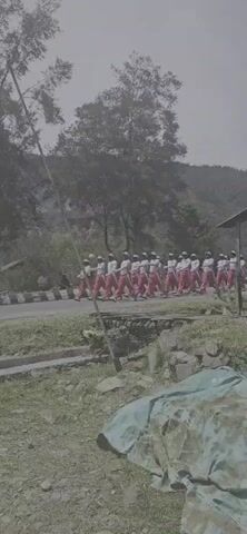 A group of people wearing white shirts and red pants are marching in formation on a paved road. They are wearing hats. A flag, which appears to be the Indonesian flag, is visible on a pole to the left of the group. The background consists of trees and a hillside. The weather appears overcast. There is some greenery on the ground in the foreground.