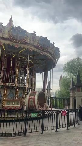 This media file captures a view of a carousel in an amusement park, with a castle-like structure and trees in the background. The carousel is ornately decorated with intricate designs, ornate carvings, and colorful lights. Two young girls are seen riding on horses on the carousel, appearing to be enjoying their ride. The sky is cloudy, suggesting a possibly overcast day, though the overall lighting is bright enough to clearly see the details of the carousel and its surroundings. A fence in the foreground separates the viewer from the attraction, and a green sign with the word "ВЫХОД" (Exit in Russian) is visible on the carousel structure. The setting appears to be a recreational area, possibly a park or amusement park, in Moscow, Russia, given the context. The mood conveyed is one of simple childhood enjoyment and a whimsical atmosphere.