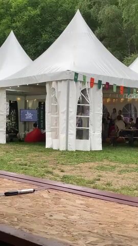 The video shows a group of people at an outdoor event. White tents are set up on a grassy area, with trees in the background. Some people are inside the tents, while others are outside, walking around or socializing. There's music playing in the background. A wooden table is visible in the foreground.