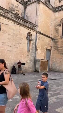 A young boy in a blue t-shirt drinks from a water bottle, walking in an outdoor courtyard paved with grey and brown tiles. Behind him, a man in a brown t-shirt and shorts stands near a speaker and a music stand. The man appears to be playing music, possibly singing or speaking. The background features a large, old building constructed of light-colored stone blocks, with arched windows and ornate architectural details. The sky is a clear blue. The camera pans upwards to show more of the building's facade and the sky. The video appears to be shot during daylight.