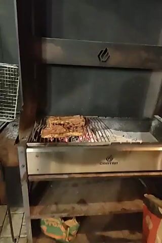 The video shows a grill with meat cooking over coals. The grill appears to be made of metal and has a branded logo. To the right of the grill, there is a wooden gazebo with wooden benches and a table. On the table, there are bottles, paper towels, and a basket. The gazebo is decorated with fairy lights. The scene suggests an outdoor gathering or event.