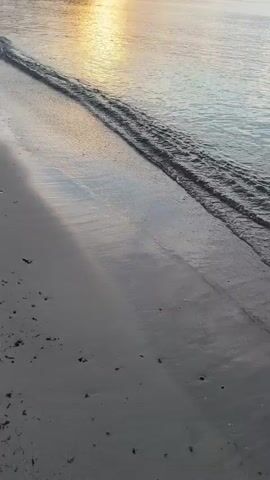 The video shows a beach scene at sunset or sunrise. The camera is positioned low to the ground, facing a body of water. Gentle waves are lapping onto a sandy shore. The sun is visible in the distance, reflecting off the water and casting a warm glow. The sky is filled with scattered clouds, some of which are illuminated by the sun. There is a faint silhouette of trees and buildings on the far shore, as well as some rocky outcrops in the water. The overall impression is one of tranquility and natural beauty. There are no people or animals visible in the video.