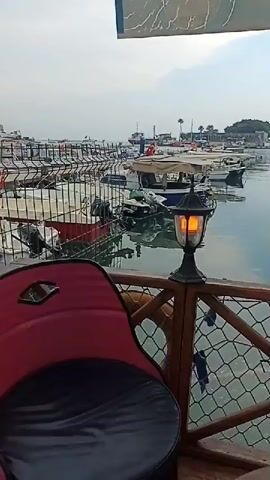 A video shows a close-up of a lit lantern on a wooden railing overlooking a harbor. Boats are docked in the background, with a fence in the foreground. A red chair is partially visible to the left. The sky is overcast. The lantern is black with a glass shade containing an orange, illuminated element. The railing has a mesh-like material attached to it. The overall scene suggests a calm, possibly early morning or evening ambiance by the water.