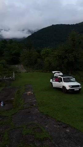 A drone pans over a rural mountain landscape. In the foreground, a white SUV is parked on a grassy clearing, its trunk open, revealing camping gear. To the left, a disused concrete structure is visible with some items on it. Further back, trees and vegetation cover the slopes of the mountains. In the distance, clouds drift across the sky, partially obscuring the mountain peaks. The scene appears to be set during either sunrise or sunset, with soft, diffused light.