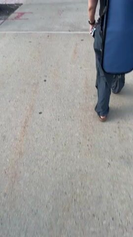 The video shows a white autonomous delivery robot moving along a sidewalk. The robot has a screen displaying two yellow triangles, a cylindrical sensor on top, and a pink flag. A person's arm, wearing a watch, enters the frame on the right side. In the background, a street with traffic and a bike lane is visible. A cyclist on a bike with a pink basket is also seen. The robot has "AVRIDE" and "Uber Eats" logos on its side. The sidewalk is made of concrete with some red markings.