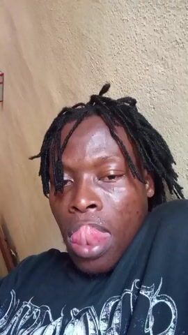 A close-up, handheld video shows a man with dreadlocks. He is wearing a black shirt with a white design. His face appears shiny and sweaty. Initially, he sticks his tongue out slightly, then pulls it back in. He then looks at the camera. The background is a beige wall. A wooden chair and other objects are visible behind him, slightly out of focus.