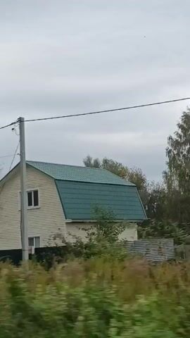 This is a video taken from a moving vehicle, likely a car, showing houses and trees passing by. The sky is overcast. There are power lines visible in the foreground. The houses appear to be in a rural or suburban setting, with fences and some vegetation. One house is painted red with a red roof, and another is light-colored with a green roof. There are also glimpses of trees with green and yellow leaves, suggesting it may be autumn. The overall impression is of a drive through a residential area on a cloudy day.